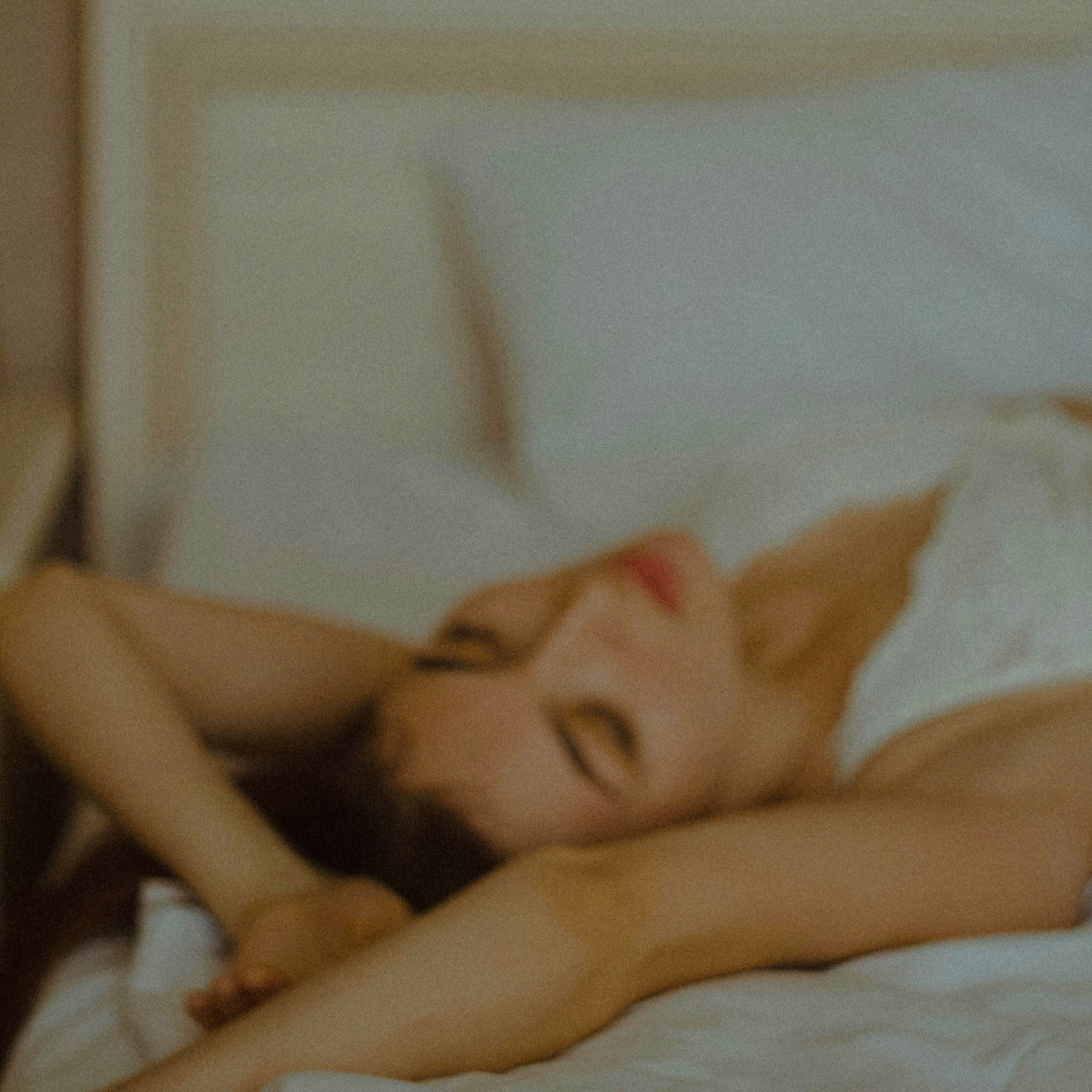 A dreamy, soft-focus close-up of a woman’s face as she rests on bed sheets, captured with a vintage film grain and warm, intimate lighting.